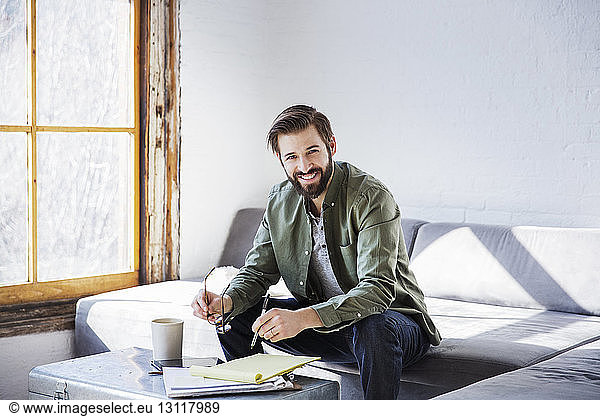 Portrait of smiling businessman writing in book while sitting in creative office