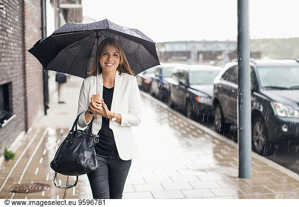 Portrait of smiling businesswoman walking on sidewalk with umbrella during rainy season