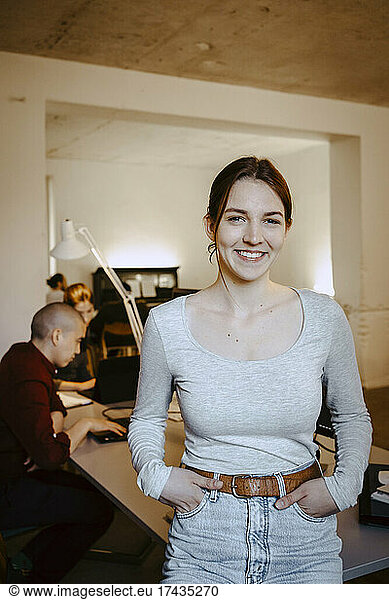 Portrait of smiling businesswoman with hands in pockets while colleague working at desk in office