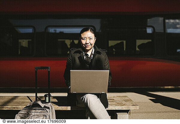 Portrait of smiling businesswoman with laptop sitting on bench in front of train at railroad station