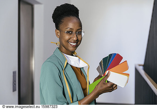 Portrait of smiling businesswoman with protective mask holding color sample in staircase