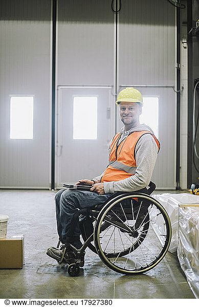 Portrait of smiling carpenter with disability holding digital tablet while sitting on wheelchair in warehouse