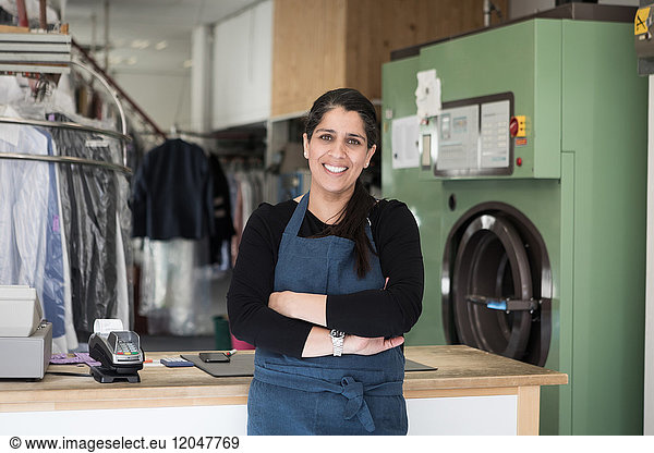 Portrait of smiling confident mature female dry cleaner standing with arms crossed at Laundromat