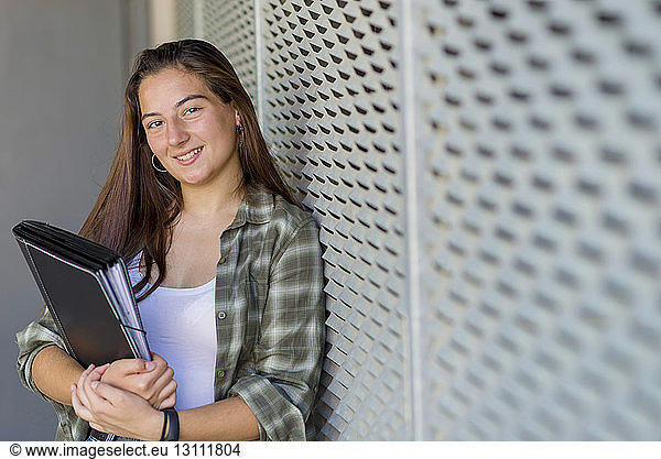 Portrait of smiling confident student holding file while standing against patterned wall in city