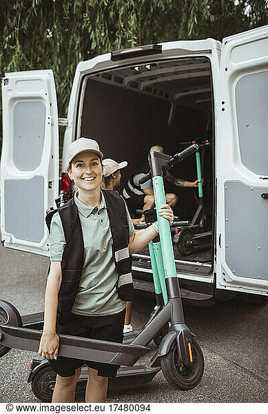 Portrait of smiling delivery woman carrying electric push scooter against coworkers