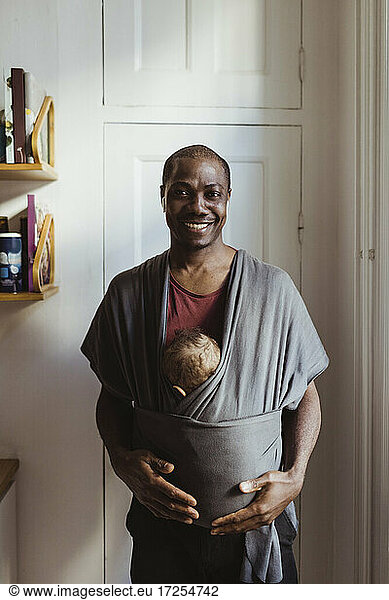Portrait of smiling father carrying son in baby carrier at home