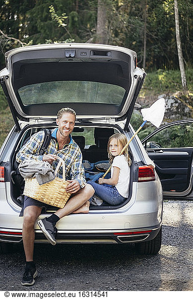 Portrait of smiling father with picnic basket by daughter sitting in car trunk