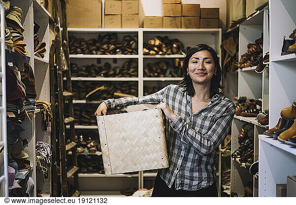 Portrait of smiling female clerk carrying wicker basket while standing amidst shoe racks at store
