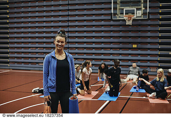 Portrait of smiling female coach standing with exercise mat against students in basketball court