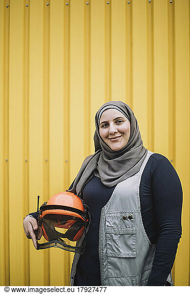 Portrait of smiling female construction worker in headscarf standing with hardhat against yellow metal wall