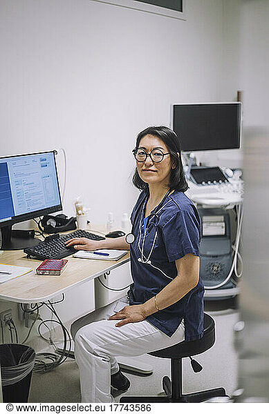 Portrait of smiling female doctor wearing eyeglasses sitting at desk in medical clinic