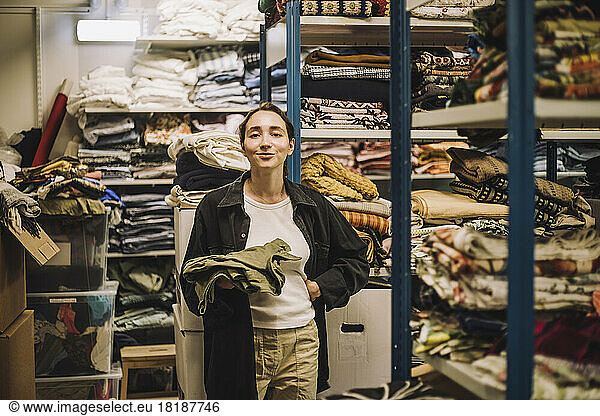 Portrait of smiling female fashion designer with hand on hip standing near clothes rack at workshop
