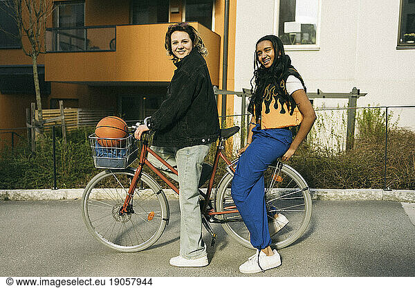 Portrait of smiling female friends sitting on bicycle in front of building at footpath