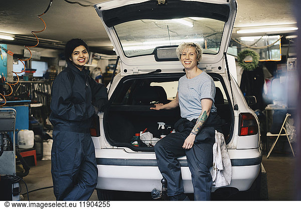 Portrait of smiling female mechanics with open car trunk at auto repair shop