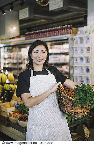 Portrait of smiling female retail clerk carrying wicker basket while standing at organic grocery store