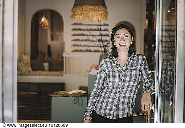 Portrait of smiling female sales clerk leaning on fashion boutique door handle