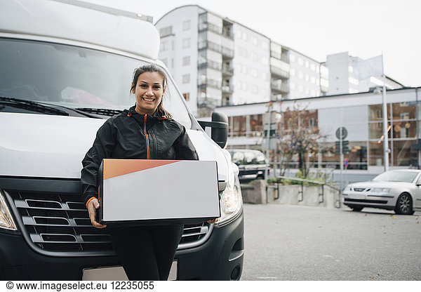 Portrait of smiling female worker carrying box while standing against delivery van in city