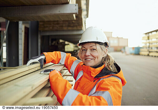 Portrait of smiling female worker in protective workwear stacking planks at industry