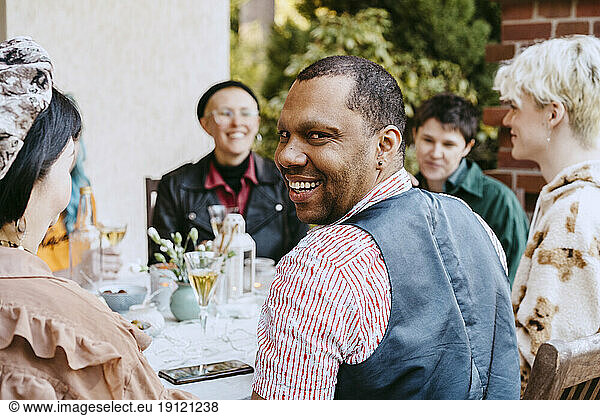 Portrait of smiling gay man looking over shoulder by friends during dinner party in back yard