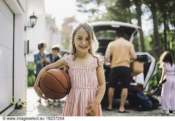 Portrait of smiling girl holding basketball with family in background
