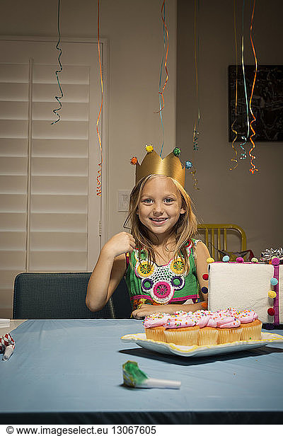 Portrait of smiling girl sitting by table with birthday present and cup cakes at home