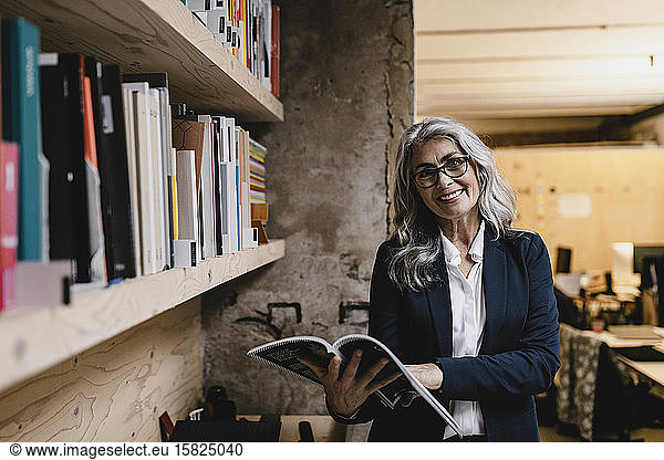 Portrait of smiling grey-haired businesswoman holding a magazine in a loft office