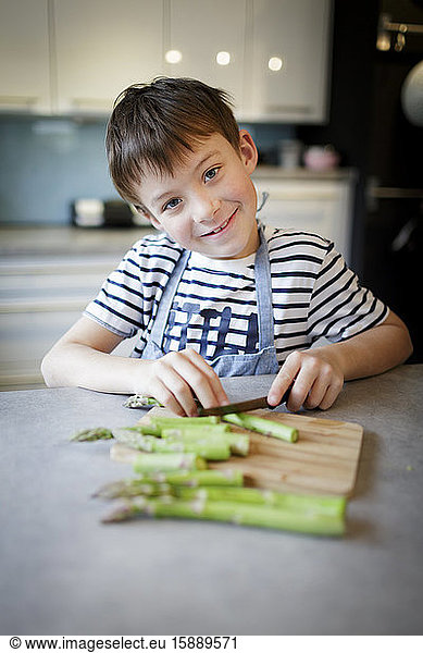 Portrait of smiling little boy cutting green asparagus in the kitchen