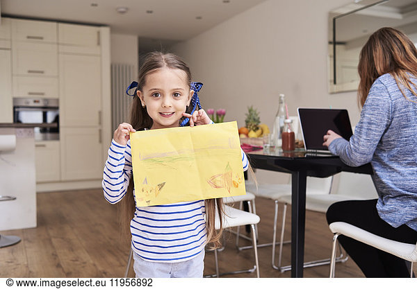 Portrait of smiling little girl showing drawing while her mother working on laptop in the background