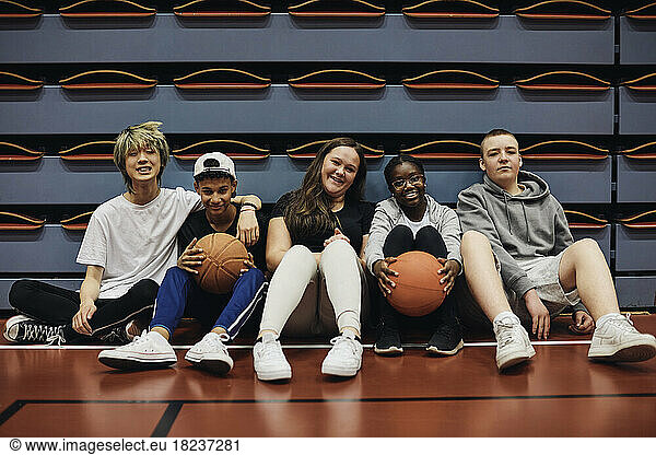 Portrait of smiling male and female friends sitting with basketballs in sports court