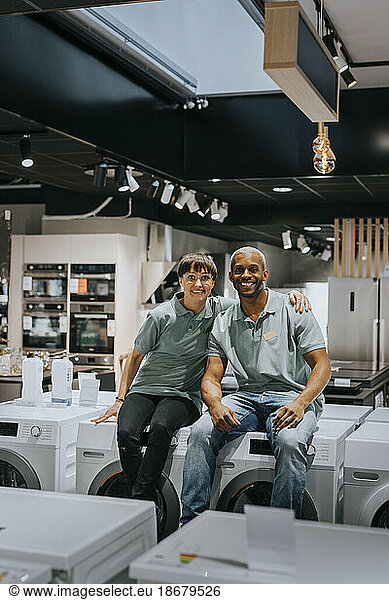 Portrait of smiling male and female retail clerks sitting on washing machines in electronics store
