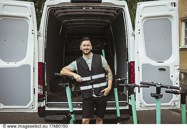 Portrait of smiling male blue-collar worker leaning on push scooter against delivery van