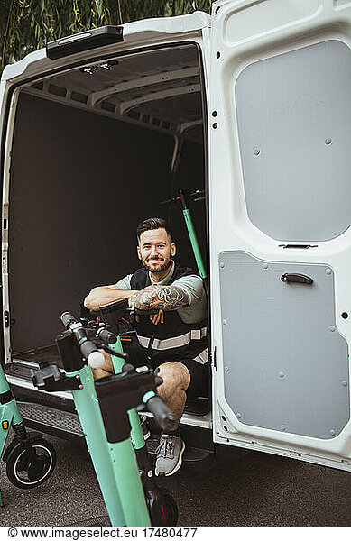 Portrait of smiling male blue-collar worker sitting in delivery van while leaning on electric push scooter