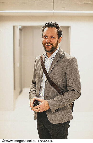 Portrait of smiling male entrepreneur with smart phone in corridor at office