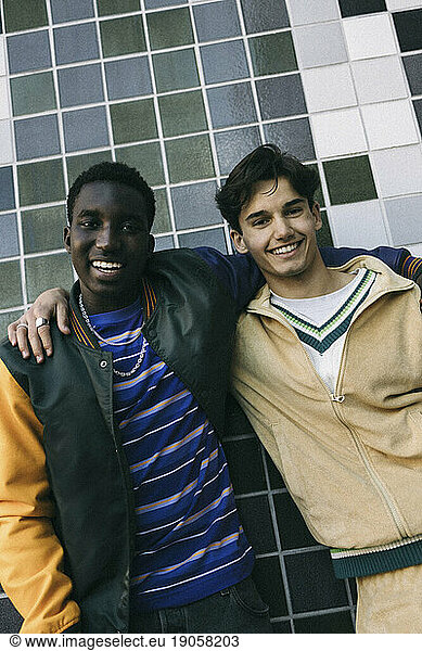 Portrait of smiling male friends standing with arms around against tiled wall
