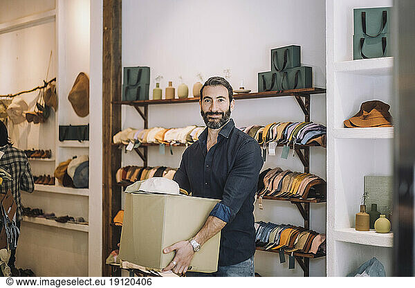 Portrait of smiling male sales clerk carrying box at fashion boutique door handle