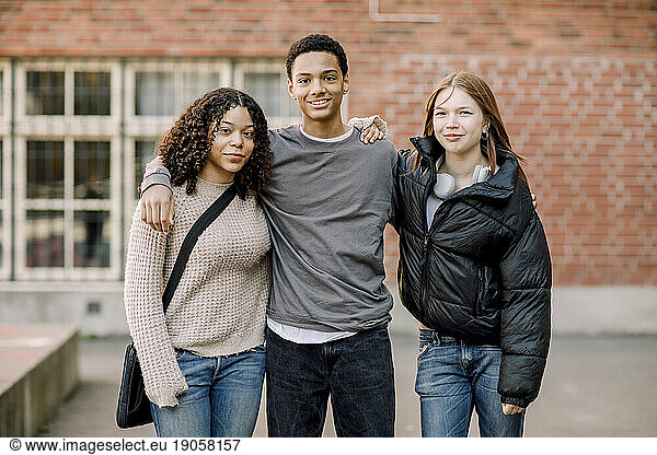 Portrait of smiling male teenage boy standing with arms around female friends in high school campus