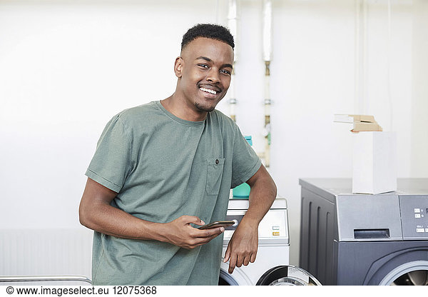 Portrait of smiling man holding mobile phone while standing by washing machine