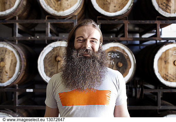 Portrait of smiling man with beard standing against barrels at brewery