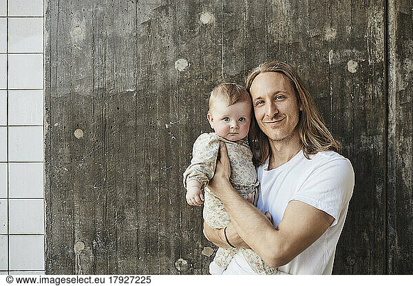 Portrait of smiling man with long hair carrying toddler daughter in front of wall