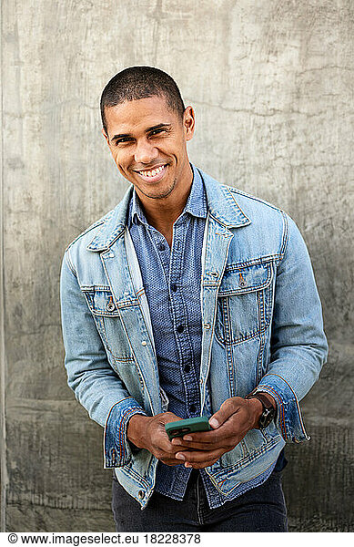 Portrait of smiling man with smart phone standing against wall