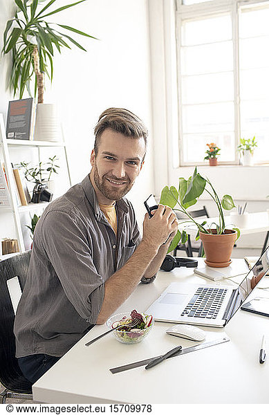 Portrait of smiling man with smartphone having lunch break at desk in office