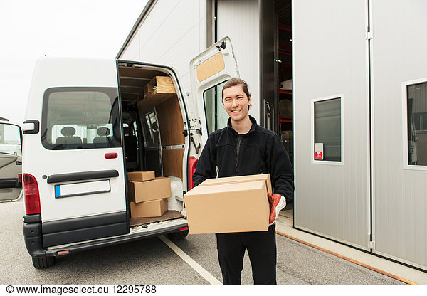Portrait of smiling manual worker carrying cardboard box while standing against delivery van