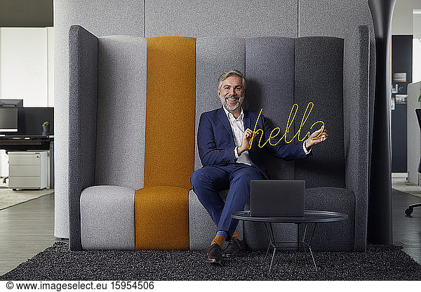 Portrait of smiling mature businessman sitting on couch in office holding hello sign