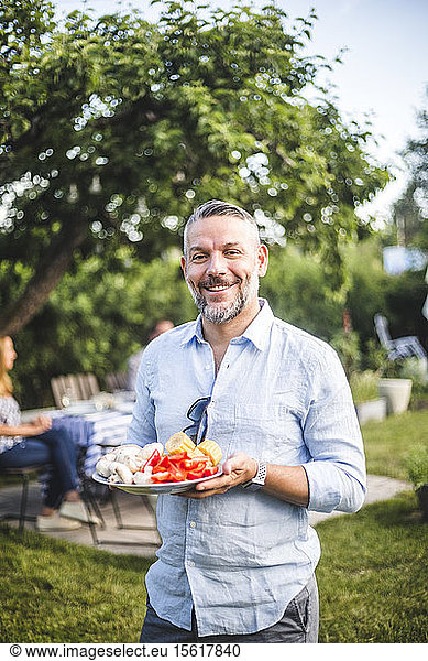 Portrait of smiling mature man holding food plate while standing in yard