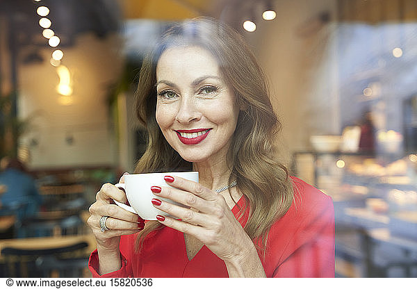 Portrait of smiling mature woman with cup of coffee behind windowpane