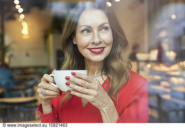Portrait of smiling mature woman with cup of coffee behind windowpane