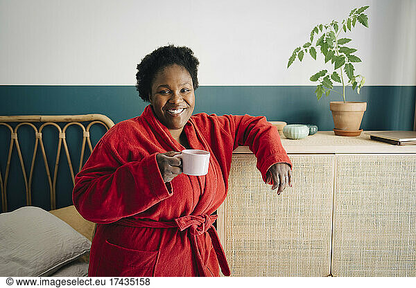 Portrait of smiling mid adult woman in read bathrobe having coffee in bedroom