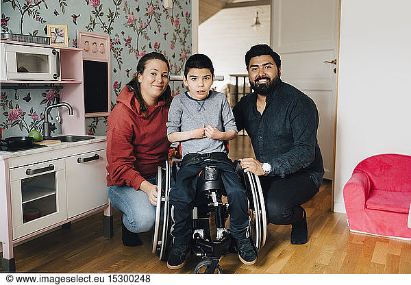 Portrait of smiling parents crouching by autistic son sitting on wheelchair at home