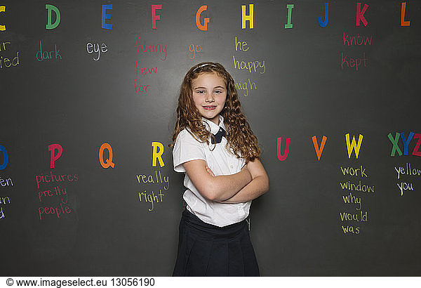 Portrait of smiling schoolgirl standing arms crossed against blackboard