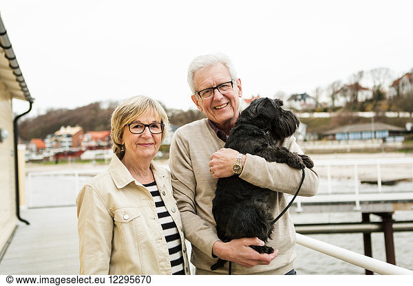 Portrait of smiling senior couple with Schnauzer at beach against sky